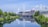 The scene features a large sports stadium with distinctive triangular white supports, located by a tranquil river. The stadium is surrounded by modern buildings and lush green trees. A bridge with multiple arches crosses over the river, and its reflection is visible on the calm water surface.