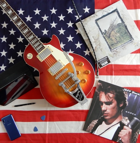 A Les Paul style electric guitar with a sunburst finish is placed on an American flag. Beside it are two vinyl records, one featuring a man holding a microphone. A pen, two guitar picks, and a smartphone accompany a tablet, all scattered on the flag.
