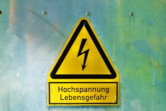 A yellow triangular warning sign with a black border and a lightning bolt symbol in the center. Below the symbol, the text reads 'Hochspannung Lebensgefahr' in black letters. The sign is mounted on a textured light blue and greenish wall with four visible screws.