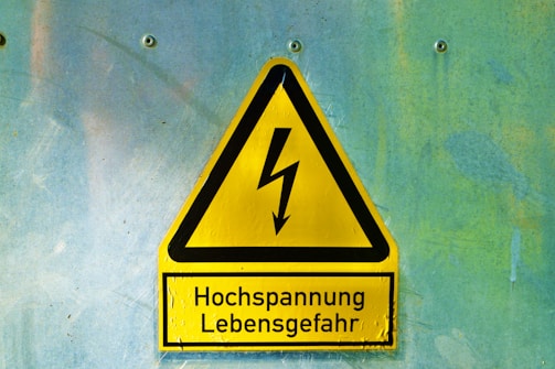 A yellow triangular warning sign with a black border and a lightning bolt symbol in the center. Below the symbol, the text reads 'Hochspannung Lebensgefahr' in black letters. The sign is mounted on a textured light blue and greenish wall with four visible screws.