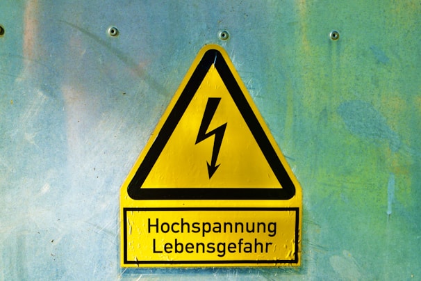 A yellow triangular warning sign with a black border and a lightning bolt symbol in the center. Below the symbol, the text reads 'Hochspannung Lebensgefahr' in black letters. The sign is mounted on a textured light blue and greenish wall with four visible screws.