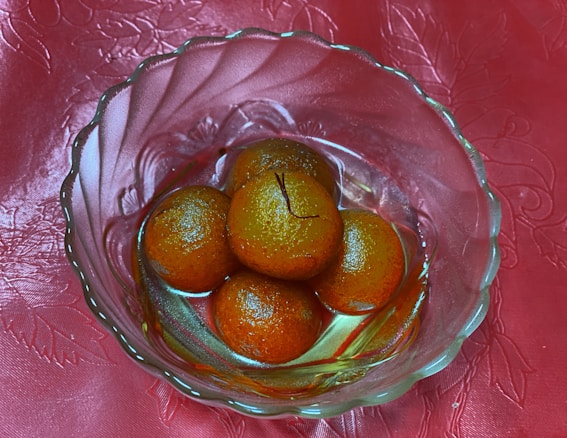 A glass bowl contains four round, brownish-orange sweets soaked in syrup, placed on a textured pink-red fabric background.