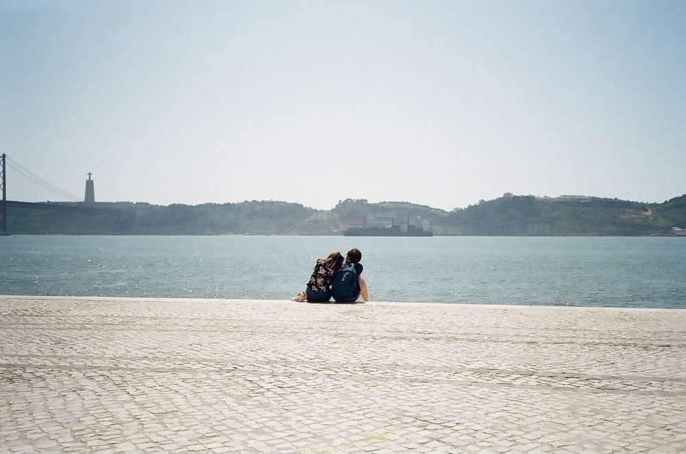 man and woman sitting on white sand near body of water during daytime