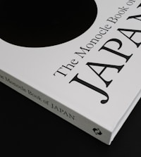 A close-up of a book titled 'The Monocle Book of Japan' with a sleek black and white cover. The book is positioned on a dark surface, creating a contrast with its white cover that features bold black text and a minimalist design.