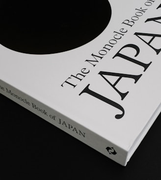 A close-up of a book titled 'The Monocle Book of Japan' with a sleek black and white cover. The book is positioned on a dark surface, creating a contrast with its white cover that features bold black text and a minimalist design.