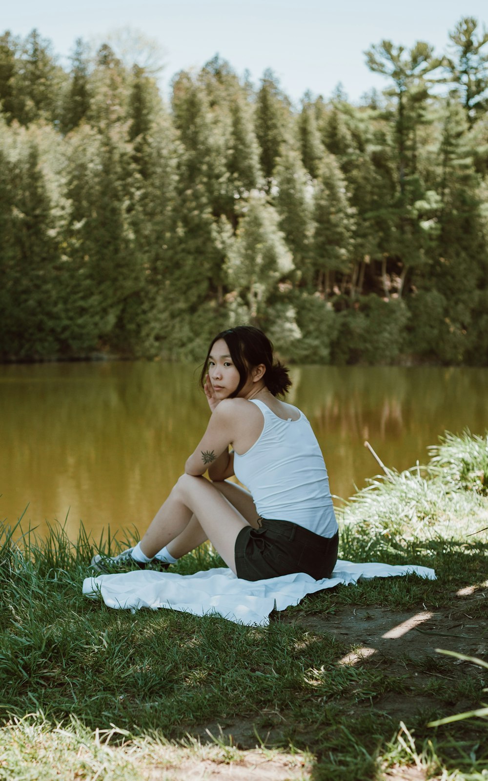 昼間、湖畔の緑の芝生に座る白いタンクトップと黒いズボンを着た女性