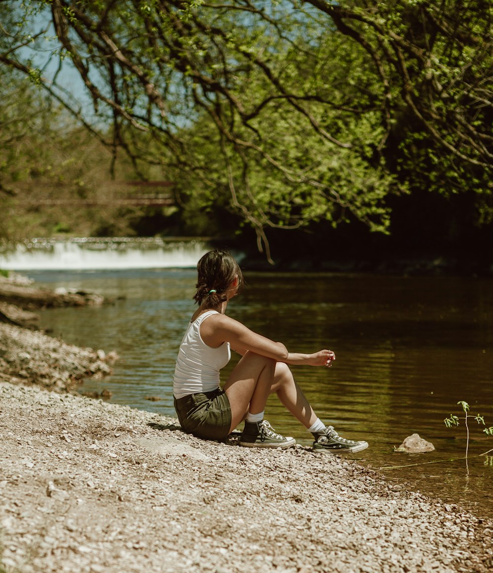 白と黒の縞模様のタンクトップを着た女の子が、昼間、川の近くの茶色の土の上に座っている