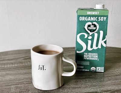 A ceramic mug filled with coffee sits on a wooden table next to a carton of Silk organic soy milk. The carton features green and white branding with text indicating it is unsweetened and contains 8g of protein per serving.