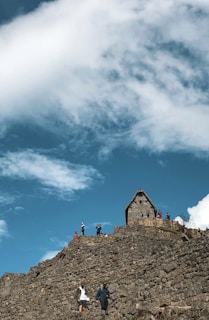A group of people is walking and standing on a stone structure, possibly ruins or a historical site, under a vibrant blue sky with scattered clouds. A small A-frame stone building is visible at the top, with several people gathered around it. The scene conveys a sense of exploration and adventure, set against a picturesque natural backdrop.