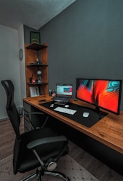 A modern workspace featuring a wooden desk with a large monitor displaying an abstract, colorful design and a laptop showing a website. The desk is equipped with a wireless keyboard and mouse on a large black desk pad. A comfortable black office chair is positioned in front of the desk. In the corner, a wooden shelf holds various items including a small speaker and decorative objects, and the wall is adorned with a circular, patterned decoration.