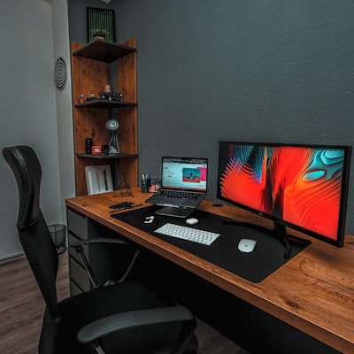 A modern workspace featuring a wooden desk with a large monitor displaying an abstract, colorful design and a laptop showing a website. The desk is equipped with a wireless keyboard and mouse on a large black desk pad. A comfortable black office chair is positioned in front of the desk. In the corner, a wooden shelf holds various items including a small speaker and decorative objects, and the wall is adorned with a circular, patterned decoration.