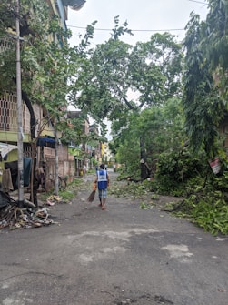A street scene after a storm with fallen branches and debris scattered on the ground. A person wearing a blue vest is walking away while holding a broom, likely engaged in cleaning efforts. Buildings line the street with partially damaged facades.