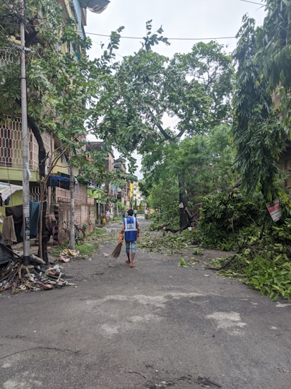 A street scene after a storm with fallen branches and debris scattered on the ground. A person wearing a blue vest is walking away while holding a broom, likely engaged in cleaning efforts. Buildings line the street with partially damaged facades.