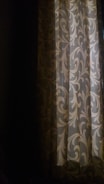 A curtain with an intricate vine-like pattern in muted shades hanging partly open, letting in a soft, ambient light. The room is mostly dark, and the light casts gentle shadows, highlighting the ornamental design on the curtain.