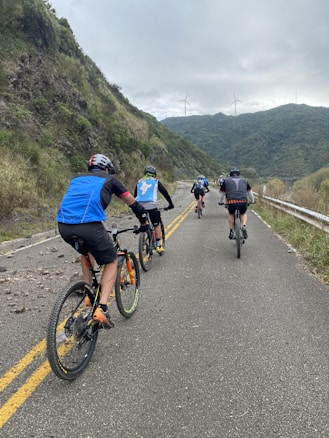 Cyclists ride along a paved road through a hilly, green landscape, with wind turbines visible on the distant hills. The sky is overcast, adding a calm atmosphere to the scene.
