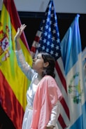 A young person dressed in a white and pink outfit is reaching upward in a pose. Behind them are national flags including those of Spain, the United States, and Guatemala, suggesting a multicultural or international event.