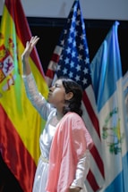 A young person dressed in a white and pink outfit is reaching upward in a pose. Behind them are national flags including those of Spain, the United States, and Guatemala, suggesting a multicultural or international event.