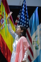 A young person dressed in a white and pink outfit is reaching upward in a pose. Behind them are national flags including those of Spain, the United States, and Guatemala, suggesting a multicultural or international event.