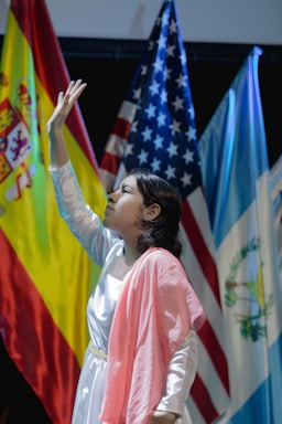 A young person dressed in a white and pink outfit is reaching upward in a pose. Behind them are national flags including those of Spain, the United States, and Guatemala, suggesting a multicultural or international event.