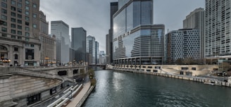 A modern urban landscape featuring tall skyscrapers positioned along a river. The water is flanked by high-rise buildings, including one with prominent glass windows and reflective surfaces. A bridge is visible in the background, and there are pathways and stairs along the riverside, suggesting pedestrian access.