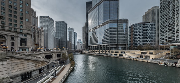 A modern urban landscape featuring tall skyscrapers positioned along a river. The water is flanked by high-rise buildings, including one with prominent glass windows and reflective surfaces. A bridge is visible in the background, and there are pathways and stairs along the riverside, suggesting pedestrian access.