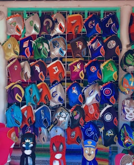 A collection of colorful luchador masks arranged on a grid display. Each mask features various designs, some with sports team logos and others inspired by superhero motifs. The masks are vibrant and include a range of colors and patterns.