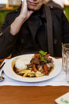 A plate of food featuring rice, grilled meat, and vegetables garnished with herbs sits on a table. The dish appears to be served with fries and some kind of sauce. A person wearing a dark jacket is seated at the table, resting their chin on their hand.