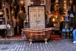 An ornate bronze bathtub is placed in front of a shop filled with intricately designed home decor items. The shop displays a variety of decorative lamps, intricate vases, and carved wooden furniture. A decorative door is centered behind the bathtub, adding to the eclectic and artisanal atmosphere.