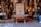 An ornate bronze bathtub is placed in front of a shop filled with intricately designed home decor items. The shop displays a variety of decorative lamps, intricate vases, and carved wooden furniture. A decorative door is centered behind the bathtub, adding to the eclectic and artisanal atmosphere.