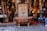An ornate bronze bathtub is placed in front of a shop filled with intricately designed home decor items. The shop displays a variety of decorative lamps, intricate vases, and carved wooden furniture. A decorative door is centered behind the bathtub, adding to the eclectic and artisanal atmosphere.