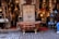 An ornate bronze bathtub is placed in front of a shop filled with intricately designed home decor items. The shop displays a variety of decorative lamps, intricate vases, and carved wooden furniture. A decorative door is centered behind the bathtub, adding to the eclectic and artisanal atmosphere.