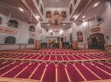 A spacious and ornate mosque interior with a rich red carpet featuring intricate patterns. The room is lit with a large, elaborate chandelier hanging from the high ceiling. Several people are standing towards the front, possibly in prayer, facing a Mihrab. The walls are adorned with Arabic calligraphy and multiple windows.