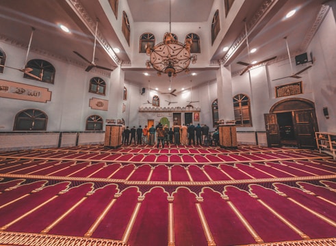 A spacious and ornate mosque interior with a rich red carpet featuring intricate patterns. The room is lit with a large, elaborate chandelier hanging from the high ceiling. Several people are standing towards the front, possibly in prayer, facing a Mihrab. The walls are adorned with Arabic calligraphy and multiple windows.