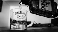 A close-up black and white photograph captures a clear glass with 'Jack Daniel's Old No. 7 Brand' logo holding some liquid while more is being poured from a bottle labeled 'Knob Creek'. The scene is set on a tabletop and in the blurred background, there is a closed book.