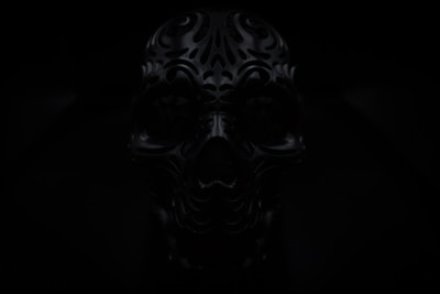 A dark image of an intricately designed, ornate skull with carved patterns. The lighting is low, adding to the mysterious and eerie atmosphere.
