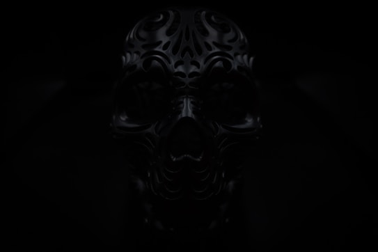 A dark image of an intricately designed, ornate skull with carved patterns. The lighting is low, adding to the mysterious and eerie atmosphere.