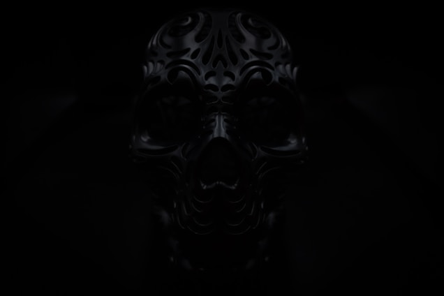 A dark image of an intricately designed, ornate skull with carved patterns. The lighting is low, adding to the mysterious and eerie atmosphere.