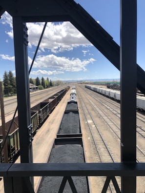 A high-angle view of a railway yard with several train tracks running parallel to each other. There is a train loaded with coal in the center track and other empty trains on adjacent tracks. The scene is framed by the structure of a bridge or an overpass, creating a rectangular border around the main view. The sky is clear with a few clouds visible, and there are trees and structures along the railway lines in the background.
