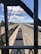A high-angle view of a railway yard with several train tracks running parallel to each other. There is a train loaded with coal in the center track and other empty trains on adjacent tracks. The scene is framed by the structure of a bridge or an overpass, creating a rectangular border around the main view. The sky is clear with a few clouds visible, and there are trees and structures along the railway lines in the background.