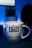A white mug with a blue and red star pattern is prominently placed in the foreground. The word 'Dad' is printed in a bold, blue font at the center, followed by the phrase 'You're a super star!' in a playful style. The background features a blurred indoor setting with a potted plant and a dark silhouette, likely a monitor or speaker.