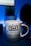 A white mug with a blue and red star pattern is prominently placed in the foreground. The word 'Dad' is printed in a bold, blue font at the center, followed by the phrase 'You're a super star!' in a playful style. The background features a blurred indoor setting with a potted plant and a dark silhouette, likely a monitor or speaker.