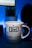 A white mug with a blue and red star pattern is prominently placed in the foreground. The word 'Dad' is printed in a bold, blue font at the center, followed by the phrase 'You're a super star!' in a playful style. The background features a blurred indoor setting with a potted plant and a dark silhouette, likely a monitor or speaker.