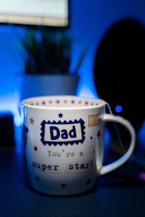 A white mug with a blue and red star pattern is prominently placed in the foreground. The word 'Dad' is printed in a bold, blue font at the center, followed by the phrase 'You're a super star!' in a playful style. The background features a blurred indoor setting with a potted plant and a dark silhouette, likely a monitor or speaker.