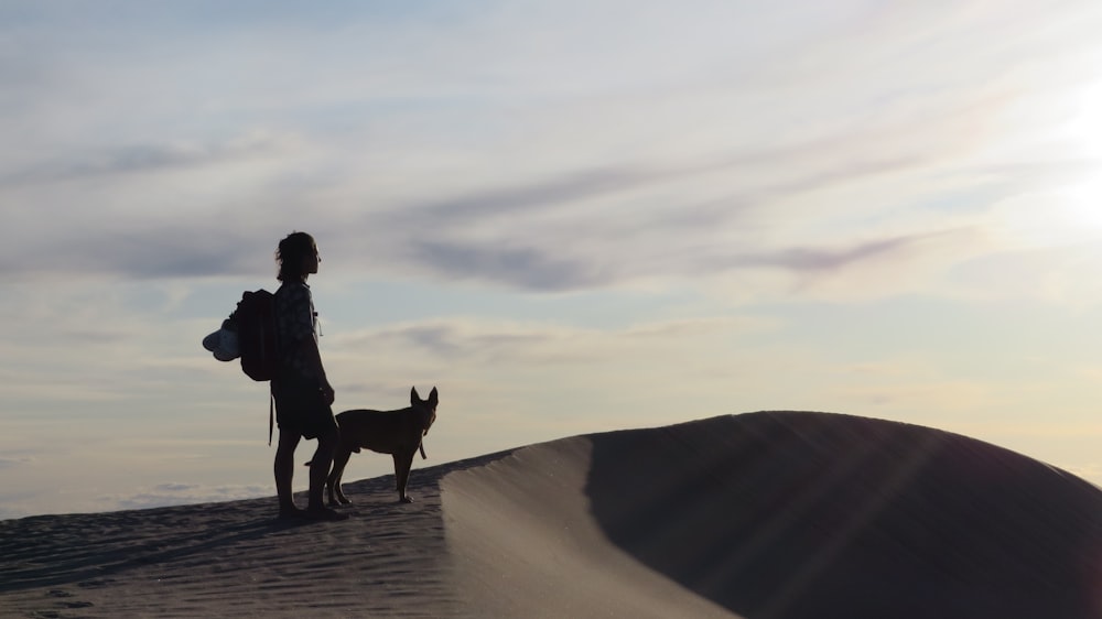 man in black jacket standing beside brown dog on brown sand during daytime