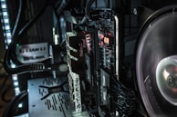 A close-up view of a computer's internal components, including a motherboard, RAM, and cooling fan. The RAM sticks have 'Trident Z Royal' branding. LED lights illuminate the area, casting a soft glow over the equipment. Various cables and connectors are neatly organized within the case.