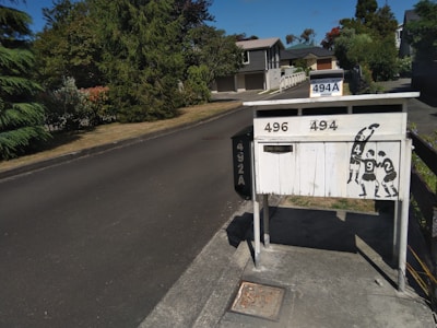 A residential street with a row of mailboxes on the right, featuring house numbers 496, 494, and 494A. The mailboxes are painted white and have a stylized black silhouette of rugby players on them. The street is lined with trees and bushes, leading to houses in the background under a clear blue sky.