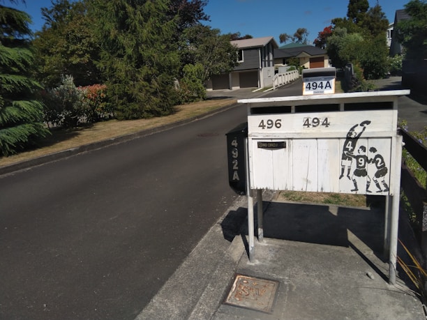 A residential street with a row of mailboxes on the right, featuring house numbers 496, 494, and 494A. The mailboxes are painted white and have a stylized black silhouette of rugby players on them. The street is lined with trees and bushes, leading to houses in the background under a clear blue sky.