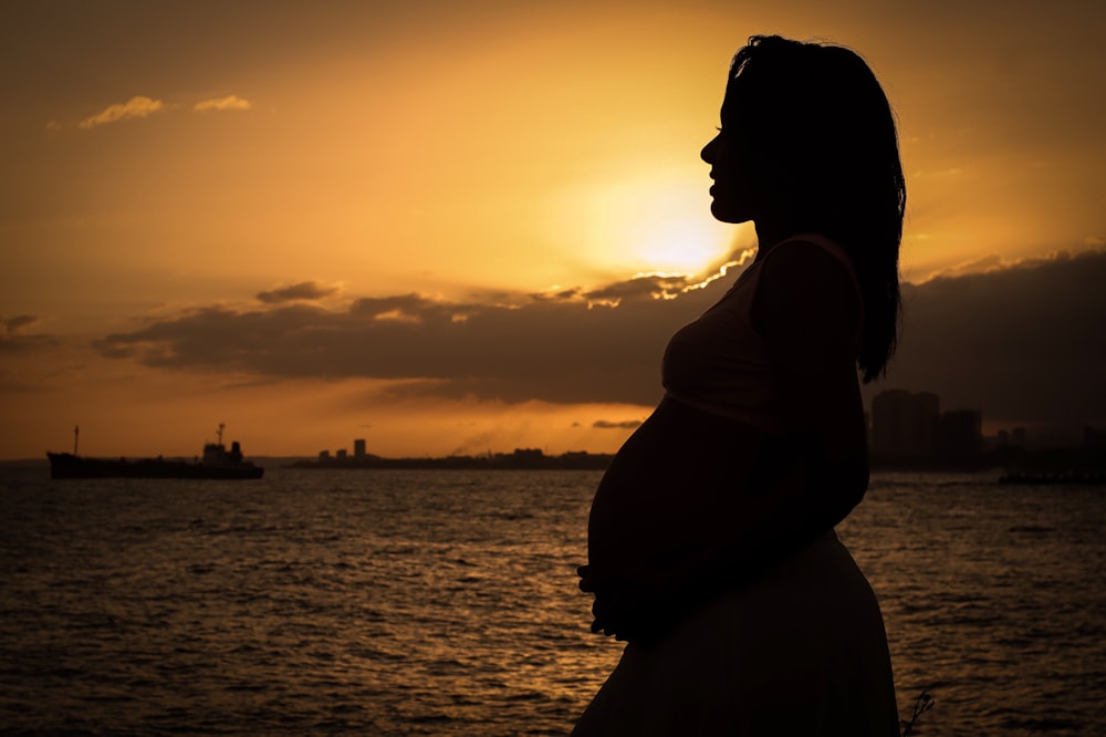 silhouette of woman standing near body of water during sunset