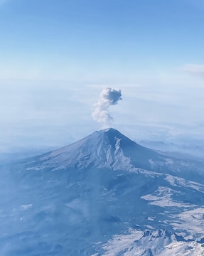 A dramatic aerial shot of a smoking volcano surrounded by green landscapes under a cloudy sky.