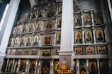 A large religious altar adorned with multiple framed icons depicting various saints and religious figures, set in a grand church interior. The icons are arranged in a structured, symmetric pattern on a multi-tiered wooden iconostasis. The walls are high and the structure has ornate decorative elements with intricate carvings.